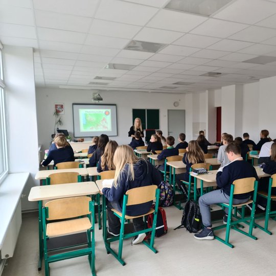 Nuotraukoje vaizduojama penktos klasės mokinių klasė. Mokiniai mokinasi temą apie Lietuvos piliakalnio kraštus. Mokytoja stovi prieš mokinius ir aiškina temą.