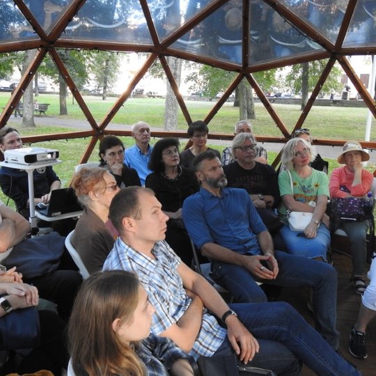 Žmonių grupė sėdi kupole, pro kupolo langus matosi parkas. Visi susikaupę klausosi pasakojimų apie archeologinius radinius.