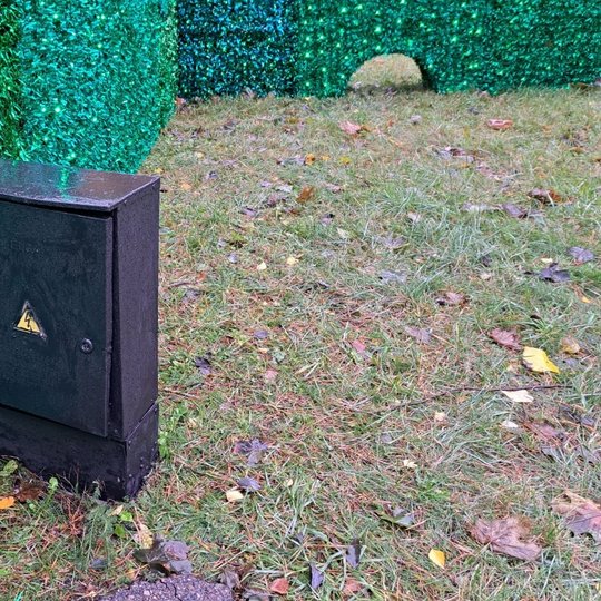 Nuotraukoje yra elektros skydinė, šviežiai nudažyta juodais dažais. Skydinė stovi ant žolės, prie žalių šviesų instaliacijos.