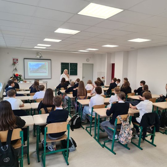 Nuotrauka vaizduoja mokinius, susėdusius prie savo klasės suolų ir klausančius mokytojos. Mokytoja klasės priekyje pasakoja vaikams pamokos temą. Įjungtas projektorius.