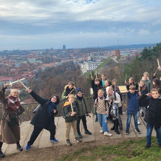 Penktokai linksmai pozuoja nuotraukai priešai Trijų Kryžių paminklą. Už nugaros matosi Gedimino pilis ir Vilniaus miesto panorama. 