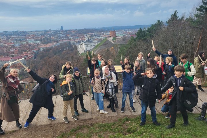 Penktokai linksmai pozuoja nuotraukai priešai Trijų Kryžių paminklą. Už nugaros matosi Gedimino pilis ir Vilniaus miesto panorama. 