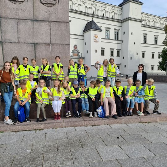 Vilniaus Filaretų pradinės mokyklos trečiokai su mokytojomis pozuoja nuotraukai prie Gedimino paminklo, esančio Katedros aikštėje. Fone matosi Valdovų rūmai ir parkas netoli rūmų ir paminklo, jame matomi esantys medžiai.