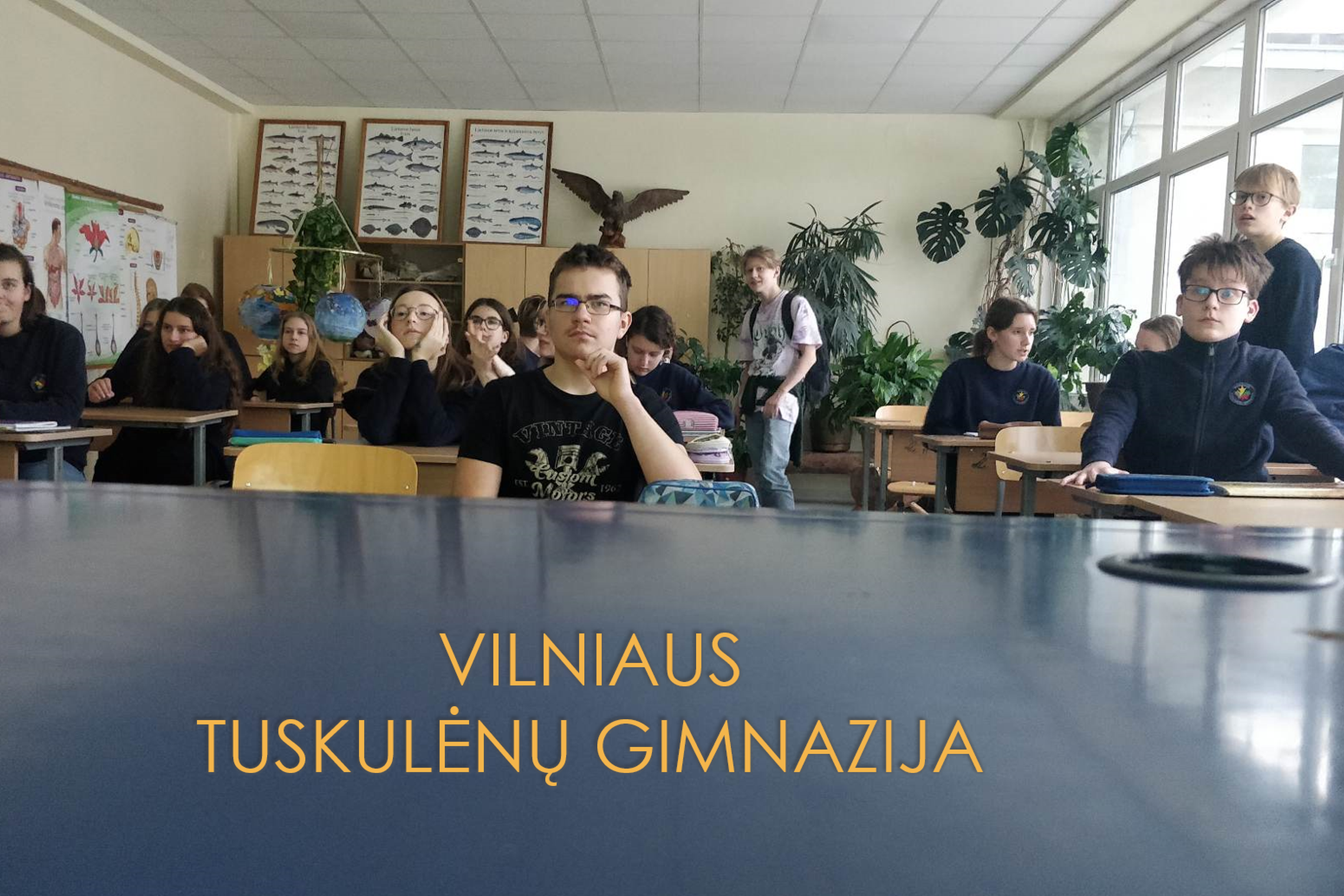 Mokiniai sėdi klasėje. Priešais nuotrauka užrašas mėlyname fone "Vilniaus Tuskulėnų Gimnazija".