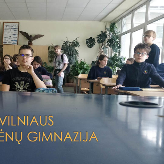 Mokiniai sėdi klasėje. Priešais nuotrauka užrašas mėlyname fone "Vilniaus Tuskulėnų Gimnazija".