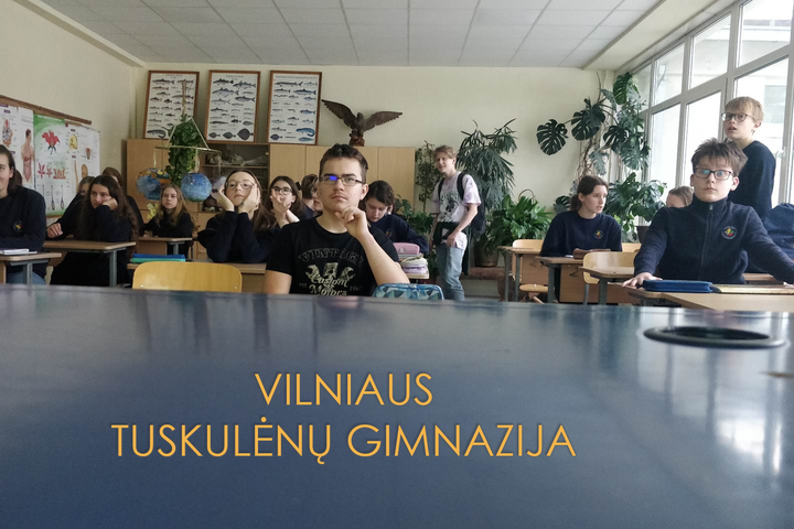 Mokiniai sėdi klasėje. Priešais nuotrauka užrašas mėlyname fone "Vilniaus Tuskulėnų Gimnazija".