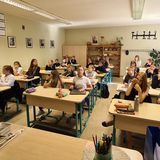 Mokiniai sėdi suoluose. Taip pat matosi mokytojos stalas, kairėje stalo pusėje yra padėtas archeologijos darbo įrankių rinkinys.  