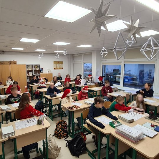 Vaikai sėdi kalėdiškai išpuoštoje klasėje, visi apsirengę kalėdiniais megztiniais.