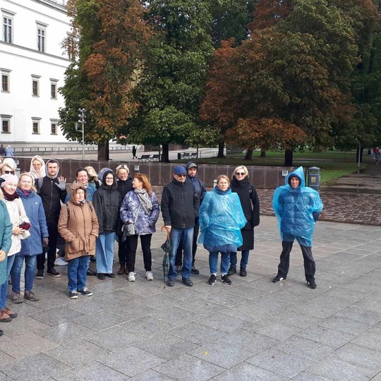 Nuotraukoje vaizduojami žmonės, kurie stovi Katedros aikštėje. Kai kurie iš jų stovi su lietpalčiais. Ekskursija vyko lietaus metu.