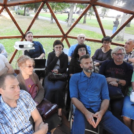 Kupole, suformuotame iš stiklinių trikampių, susispietę sėdi edukacijos dalyviai, kurie įdėmiai žvelgia į projektoriaus rodomą ekraną ir klausosi archeologinės tematikos paskaitos.