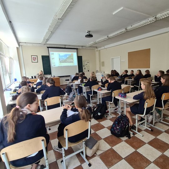 Iš klasės galo nufotografuoti klasėje sėdintys mokiniai. Mokiniai stebi mokytojos pateiktas skaidres, mokytoja sėdi prie savo rašomojo stalo.