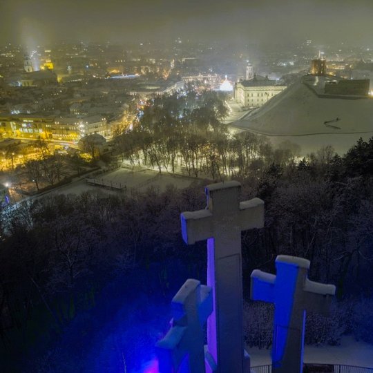Nuotrauka daryta vakare virš Trijų Kryžių paminklo. Patys Trys Kryžiai apšviesti mėlyna spalva. Tolumoje matosi Gedimino paminklas ir Sereikiškių- pilių parkas ir katedros aikštė su kalėdine miesto egle.