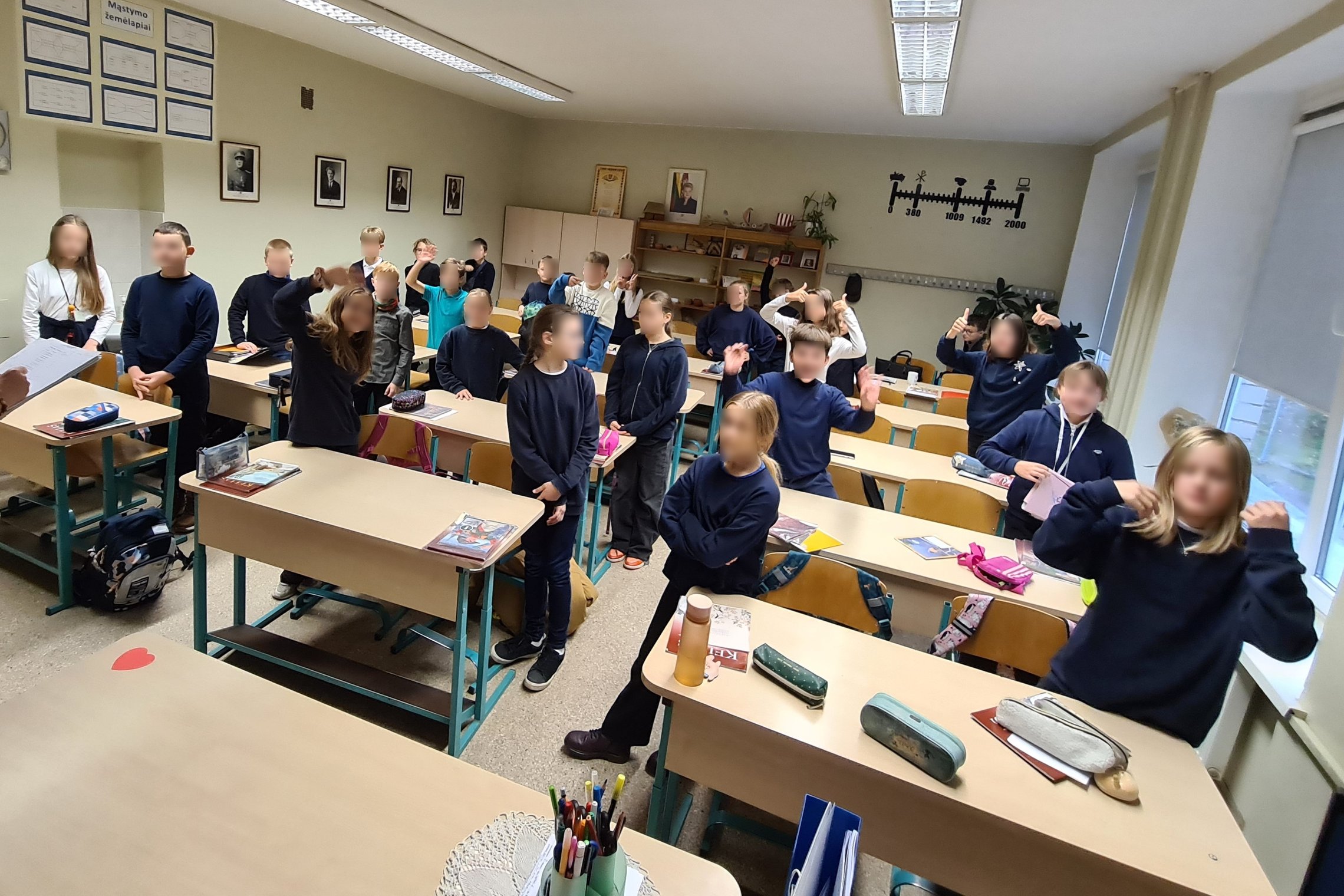 Nuotrauka vaizduoja klasėje esančius penktokus. Jie visi yra atsistoję, vieni žiūri į mokytojos pusę, kurios pilnai nesimato, o kiti pozuoja kamerai. Nuotrauka yra padaryta iš klasės priekio.