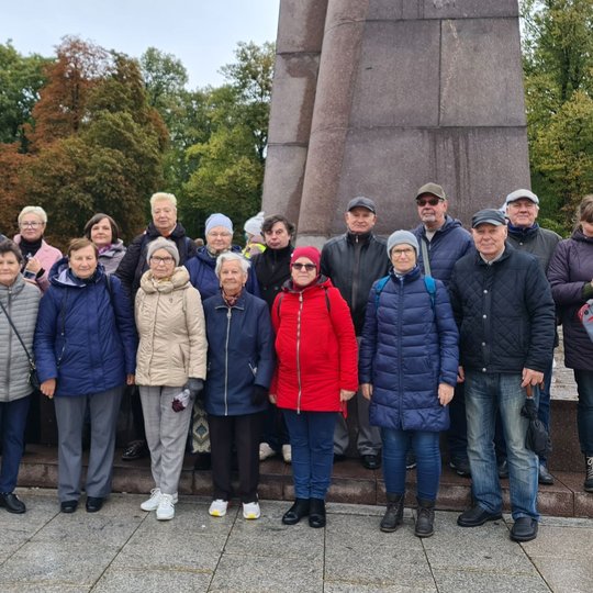 Senjorų grupelė pozuoja nuotraukai priešais Gedimino paminklą. Pilkas dangus, visi užsidėję striukes, jau šalta. Fone taip pat matosi medžiai parkelyje.