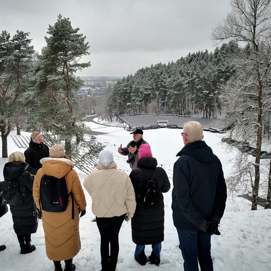Žiemišką dieną Lietuvos valstybinio simfoninio orkestro administracijos komanda klausosi edukatoriaus Stasio ant Karigailos kalvos. Tolumoje matosi Dainų Slėnio estrada.