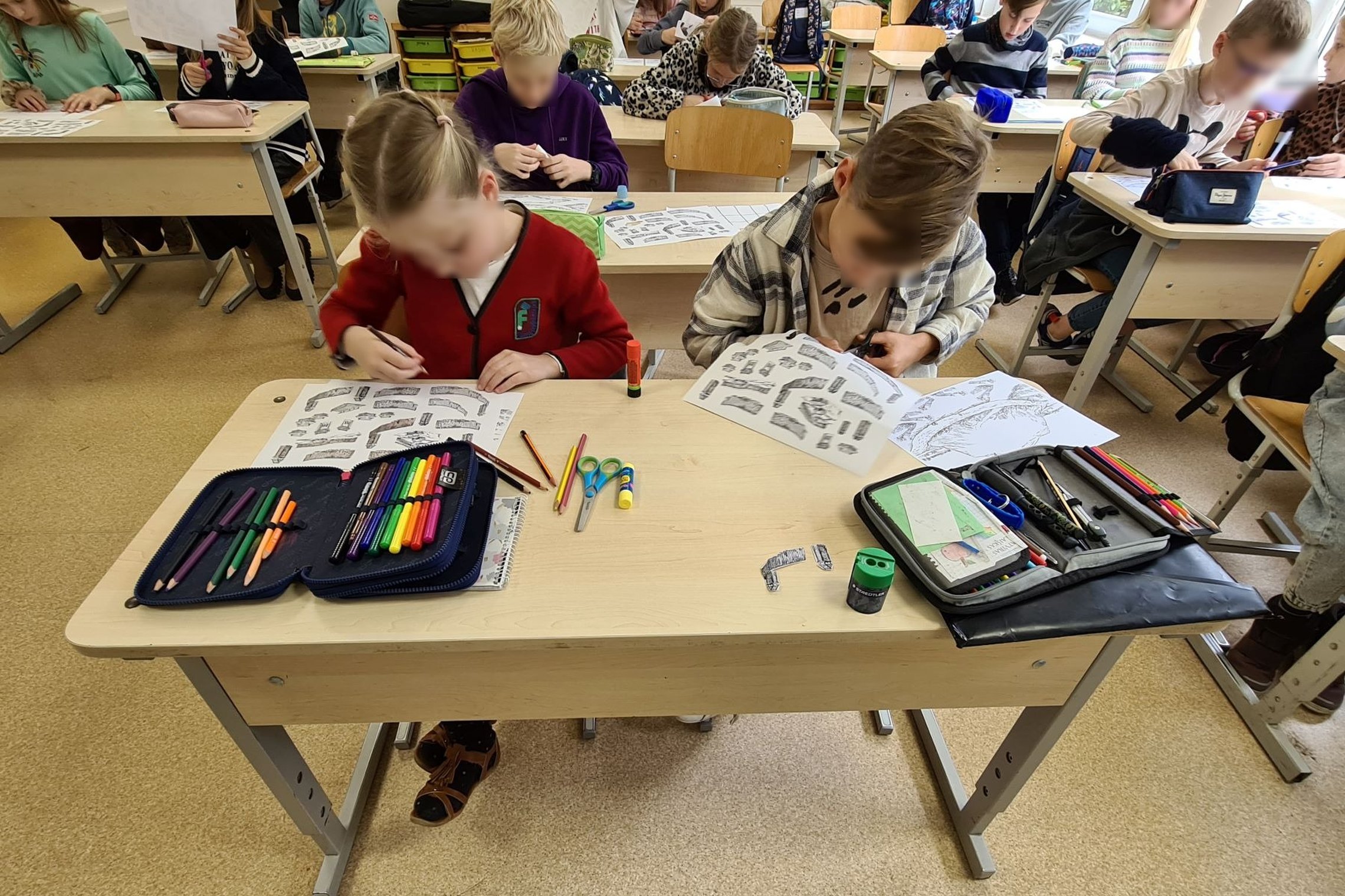 Nuotraukoje vaizduojama mokinių klasė, visiems mokiniams yra išdalinti vienodi lapai. Priekyje, pirmame suole, sėdi mergaitė ir berniukas. Mergaitė pasiėmusi pieštuką spalvina, o berniukas iš lapo bando kažką iškirpti.