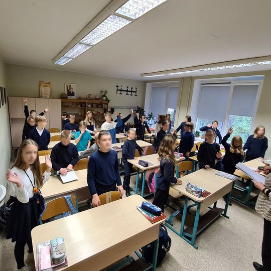 Nuotrauka vaizduoja klasėje esančius penktokus, kurie yra visi atsistoję. Dauguma iš jų mojuoja kamerai, bet kai kurie drovisi ir ramiai stovi prie savo suolų. Jų mokytoja stovi priešais klasės mokinius ir laiko kelis lapus. Nuotrauka padaryta iš klasės p