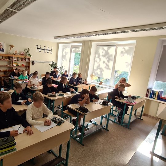 Klasėje vyksta pamoka, mokytoja pirmame suole, šalia lango, atsisėdusi prie mokinio. Visa klasė sėdi suoluose ramiai, tik kelios mergaitės gale šnekučiuojasi.