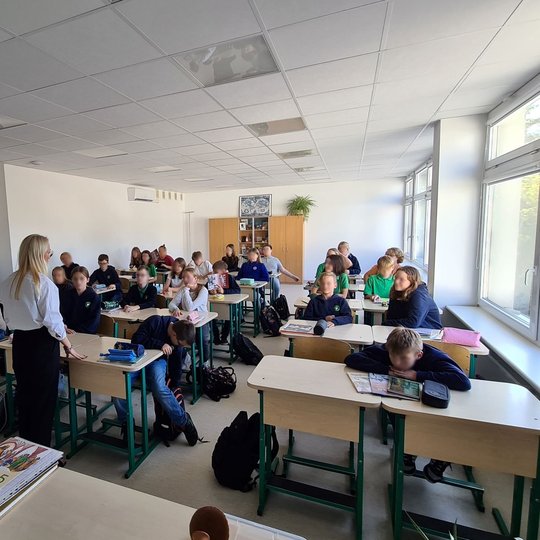 Nuotraukoje matome pilną klasę  progimnazijos mokinių. Prieš klasę stovi mokytoja, kuri aiškina temą. Mokiniai susidomėję klausosi mokytojos.
