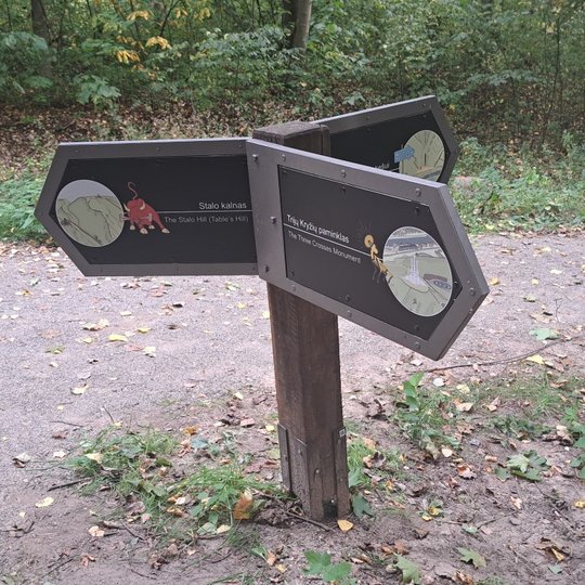 Prie keliuko stovi rodyklės, kurios rodo kryptį į tris puses, parašyti užrašai Stalo kalnas ir trijų kryžių paminklas.