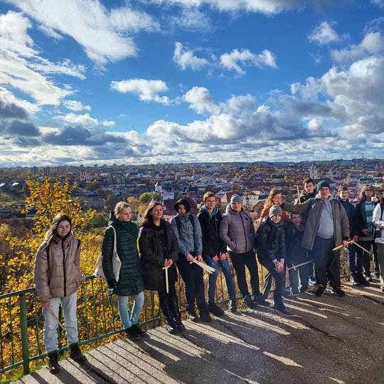 Aštuntokai pozuoja nuotraukai priešais Trijų Kryžių paminklą. Už nugaros debesuotas mėlynas dangus ir Vilniaus panorama.