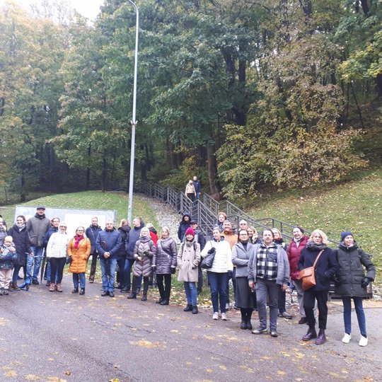 Nuotraukoje vaizduojama grupė įvairaus amžiaus žmonių. Fone, už žmonių, matosi Trijų kryžių kalnas. Du žmonės lipa laiptais nuo kalno.