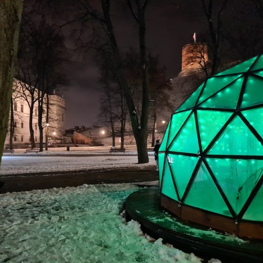 Vilniaus pilių kupolas iš lauko vakare. Tolumoje matosi Valdovų rūmų muziejus, dešinėje viršuje Gedimino kalnas su bokštu. Pats Vilniaus pilių kupolas šviečia žaliai.