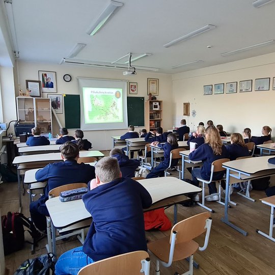 Matoma klasė mokinių, klausanti pamokos apie piliakalnių kraštą. Dauguma mokinių įdėmiai klausosi, kas yra dėstoma, o kai kurie bando užmigti beklausydami pamokos.