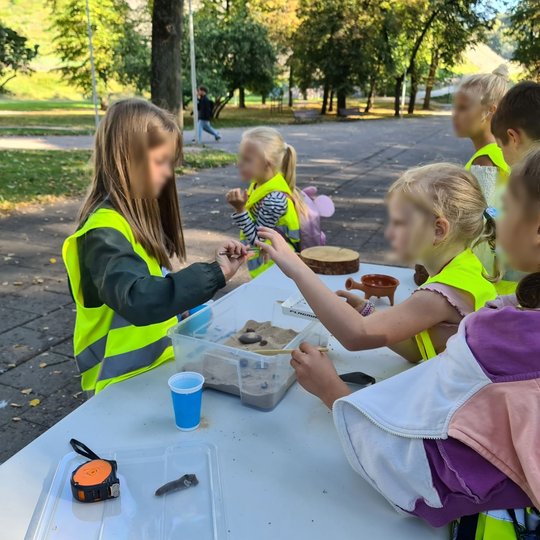 Trečiokai, vilkintys ryškias, geltonas liemenes, stovi prie stalo. Stalo viduryje pastatyta plastikinė dėžė, kurioje yra smėlio su paslėptais archeologiniais radiniais. Vaikai susidomėję tyrinėja, kas yra viduje.