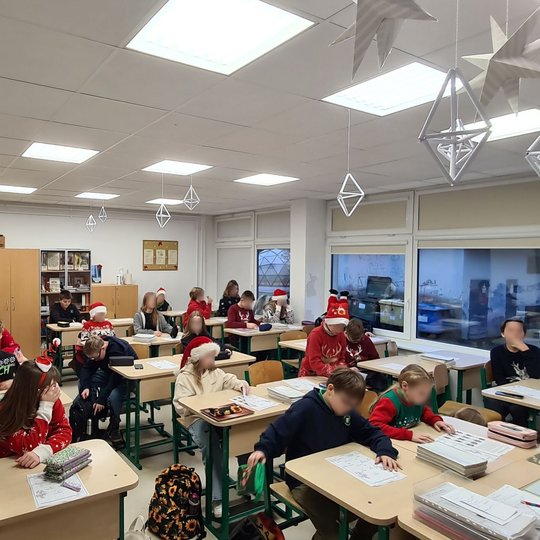 Vaikai sėdi kalėdiškai išpuoštoje klasėje, visi apsirengę kalėdiniais megztiniais.