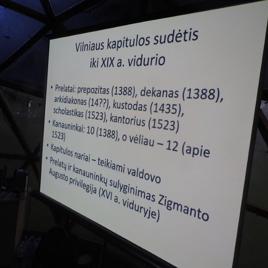 Vakaras, lauke tamsu, Vilniaus kupolo viduje yra rodoma skaidrė. Skaidrėje išsamiai aprašyta Vilniaus kapitulos sudėtis iki XIX amžiaus vidurio.
