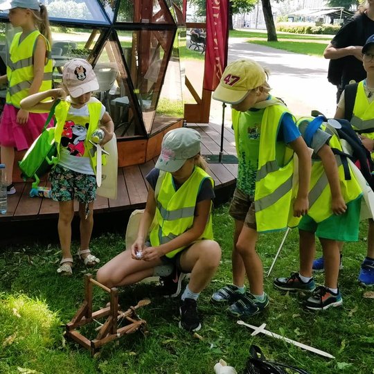 Vaikai, su šviesiai geltonomis atšvaitinėmis liemenėmis, šalia Vilniaus pilių kupolu žaidžia su katapultus modeliu.