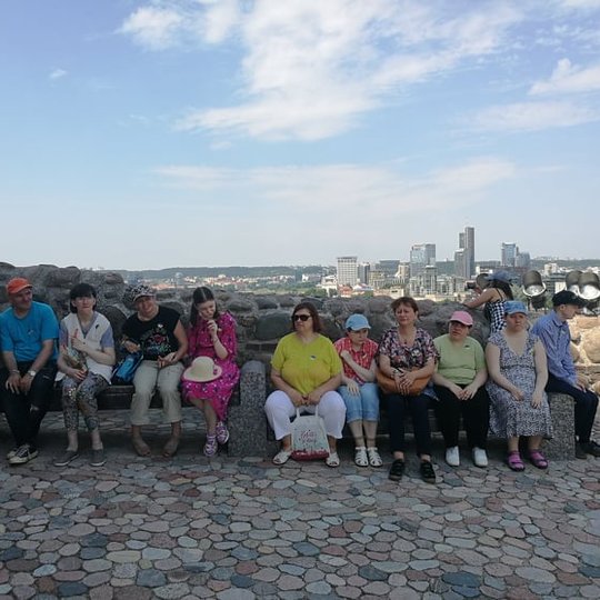 Ekskursijos dalyviai sėdi ant suoliuko. Už jų matosi Vilniaus dangoraižių panorama.