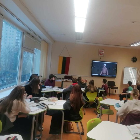 Klasėje mokiniai klausosi pamokos ir žiūri į skaidres per projektorių.