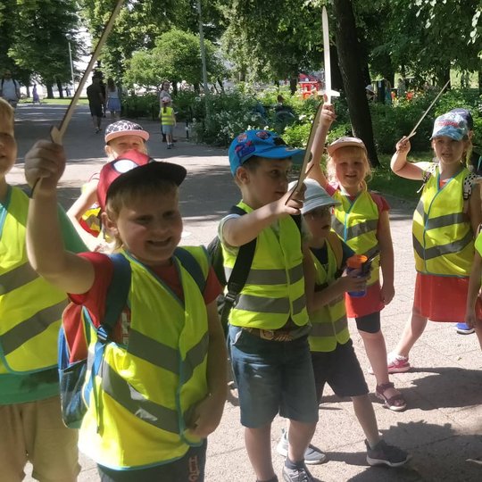 Vaikai su geltonomis atšvaitinėmis liemenėmis pozuoja iškėlę medinius kalavijus į viršų pietiniame parkelyje.