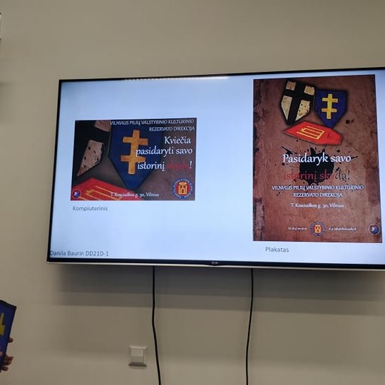 Televizoriaus ekrane rodomą skaidrė apie skydus, šalia studentas pristato savo idėją.