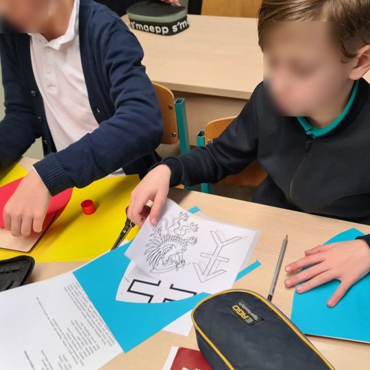 Berniukas klasės suole ruošiasi kirpti formas, tinkančias jo gaminamam herbui.