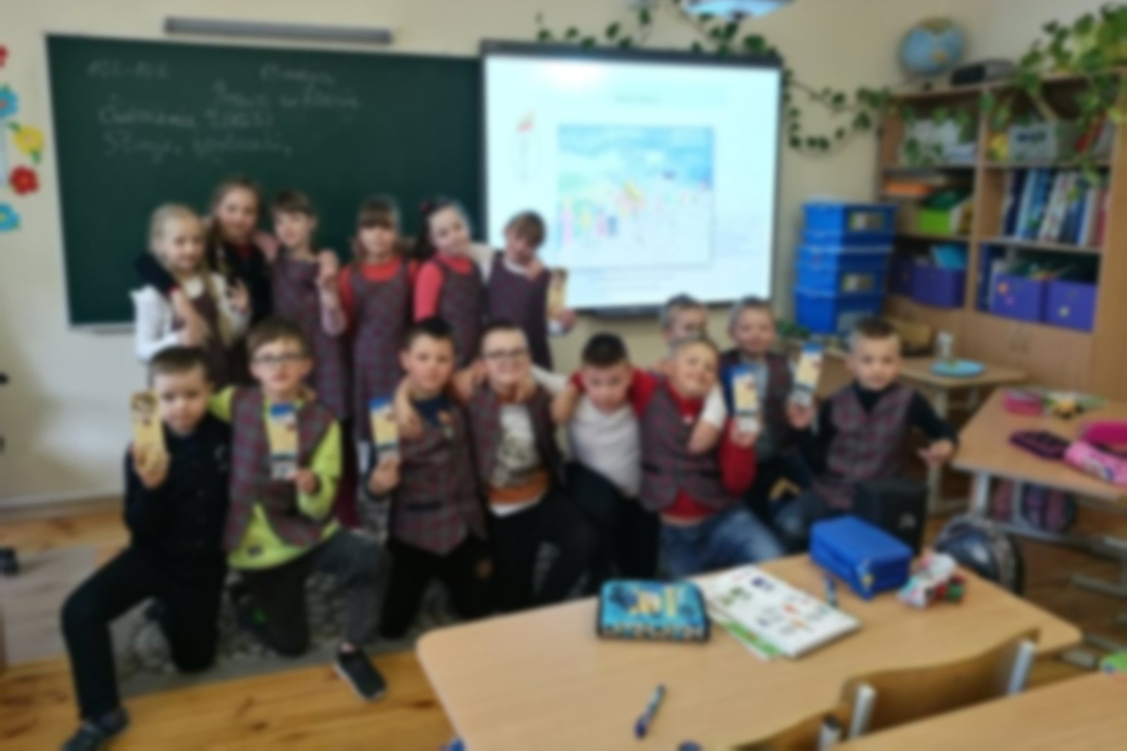 Darželinukai klasėje, jiems už nugaros lenta ir projektorius.
