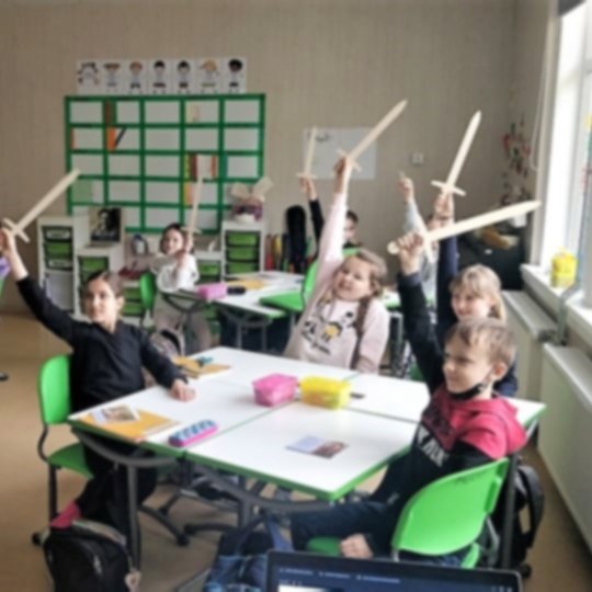 Trečiokai sėdi prie suolų klasėje, iškėlę medinius kalavijus į viršų.