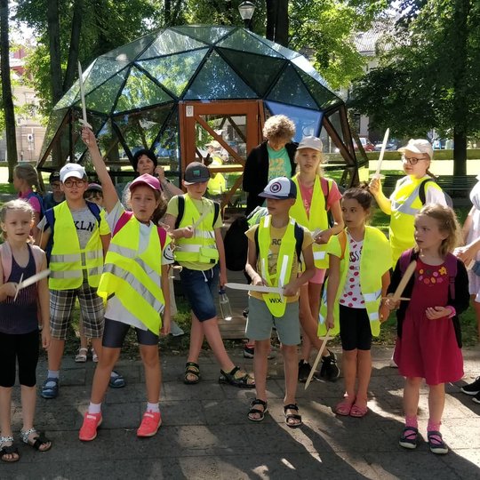 Vaikai, su atšvaitinėmis liemenėmis kaip su šarvais ir mediniais kalavijais rankose, šalia Vilniaus pilių kupolo.