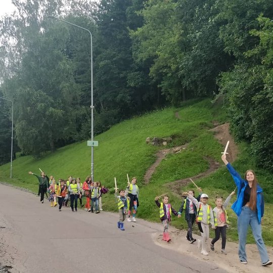 Vaikai ir auklėtojos šypsodamiesi ir iškėlę medinius kalavijus į viršų leidžiasi asfaltuotu keliu. 