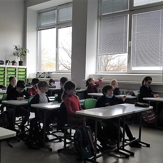 Mokiniai klasėje, sėdi prie baltų suolų, dėvi kaukes.