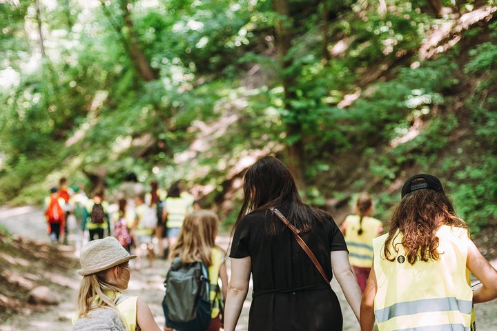 Vaikai, su geltonomis atšvaitinėmis liemenėmis ir moteris, vorele eina žaluma aplink apaugusiu taku.