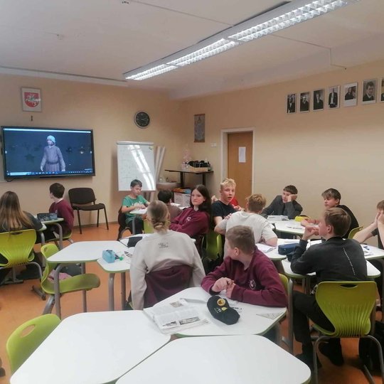 Klasėje mokiniai klausosi pamokos ir žiūri į skaidres per projektorių.