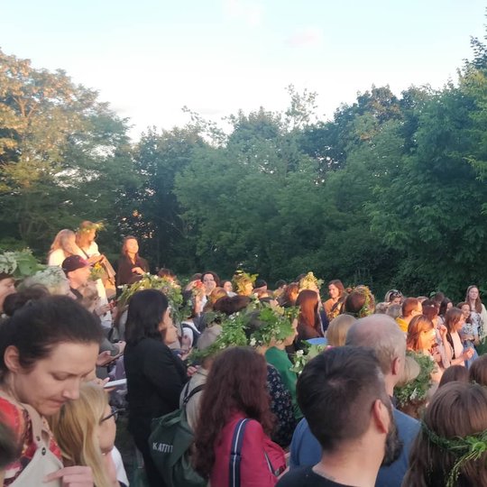 Gausi žmonių minia, kai kurie su žolių vainikais, klausosi smuikininko ir vokalistės koncerto.