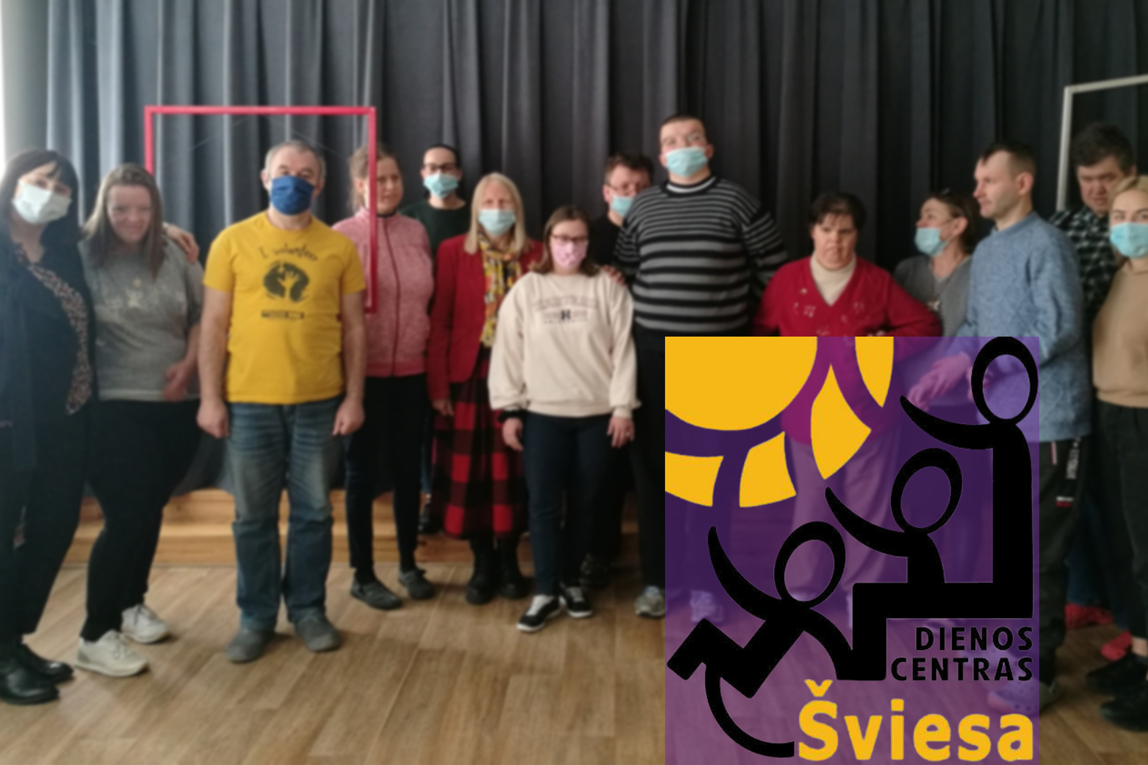 Dienos centro „Šviesa“ lankytojai pozuoja nuotraukai kartu su edukatore Angele. Nuotraukos kampe Dienos centro „Šviesa“ logotipas.