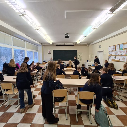 Mokiniai klasėje stovi prie suolų ir ruošiasi pamokai.
