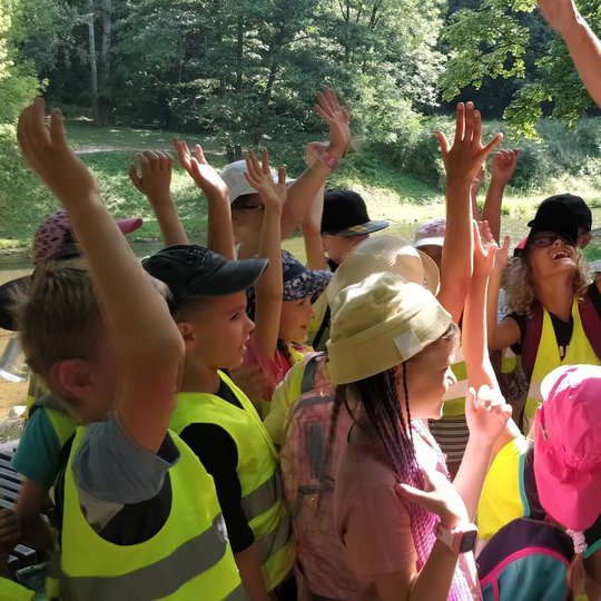 Vaikai su atšvatinėmis liemenėmis būdami būryje kelia rankas.