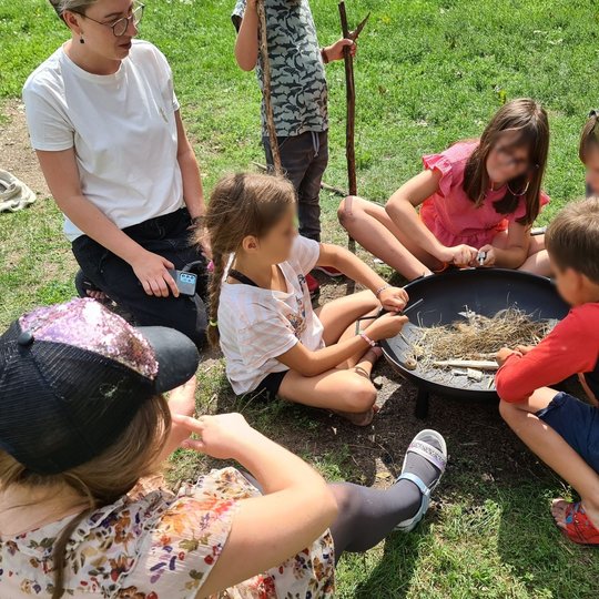 Keturių vaikų grupelė, susėdę prie laužinės, naudojasi skiltuvais.