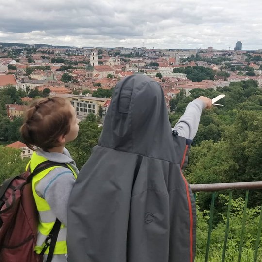 Du vaikai žiūri į Vilniaus panoramą nuo Trijų Kryžių kalno. Vaikas dešinėje, apsijuosęs galvą ir nugarą pilka striuke, rodo mediniu kalaviju į pilies skvero vakarinę dalį. 